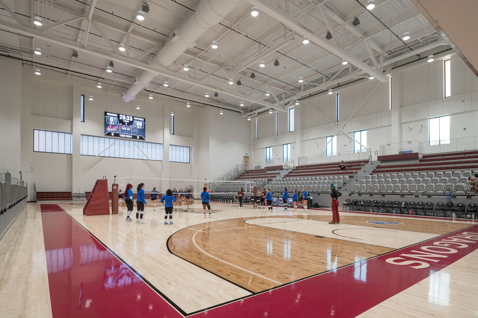 Indoor gymnasium with polished wood floor, volleyball net, and scoreboard; players warming up on...