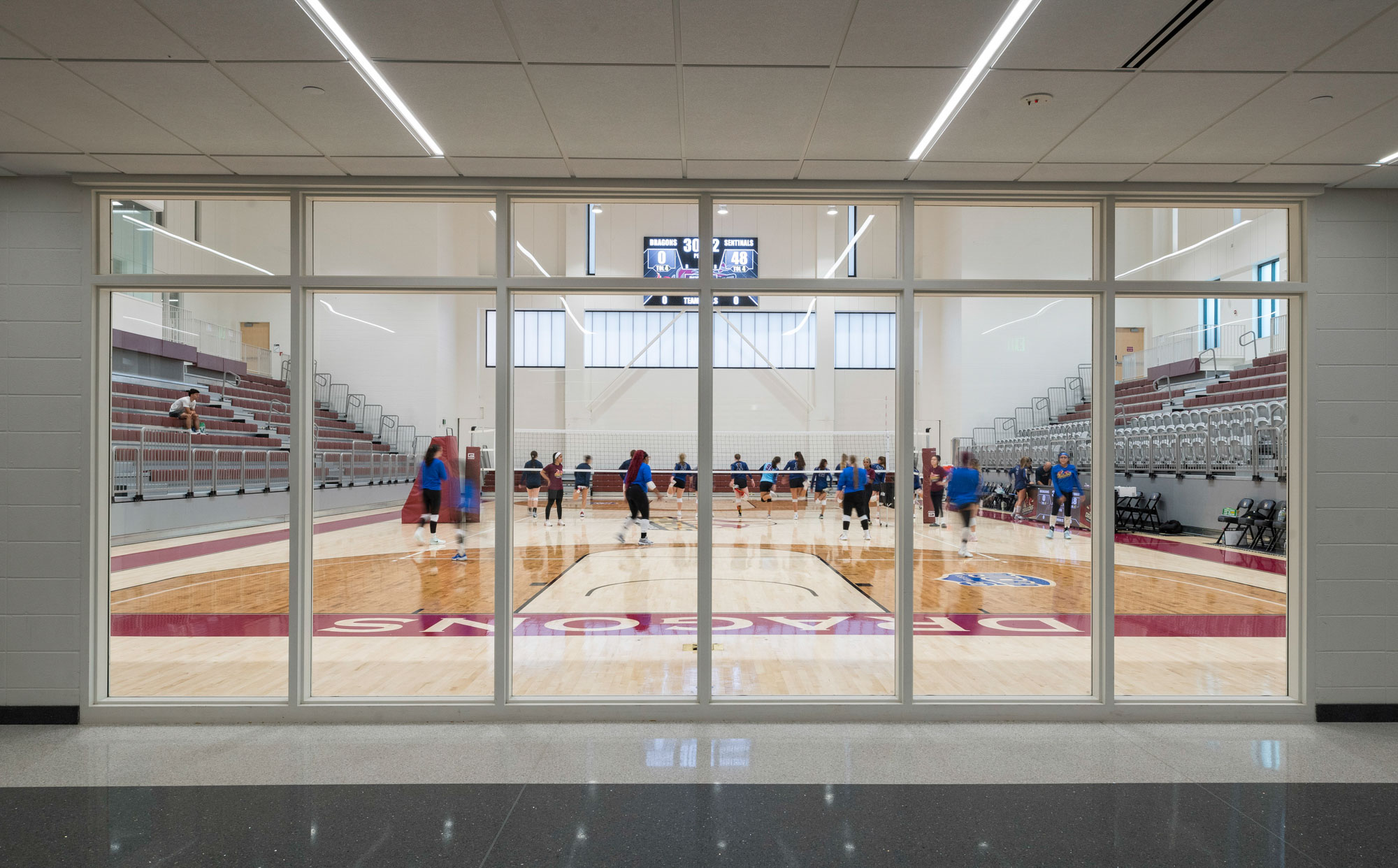 View through glass wall into gymnasium with volleyball court and seating tiers.