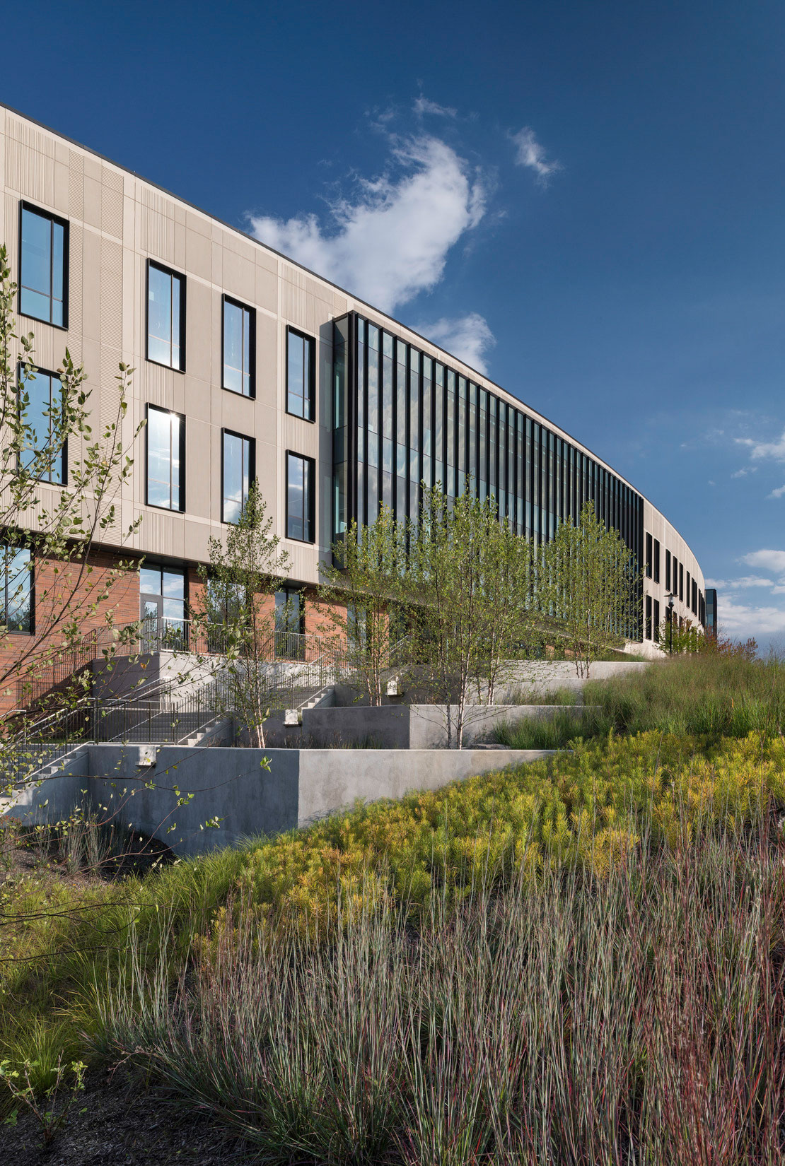 Building exterior with beige panels and brick base, framed by landscaped greenery and blue sky.