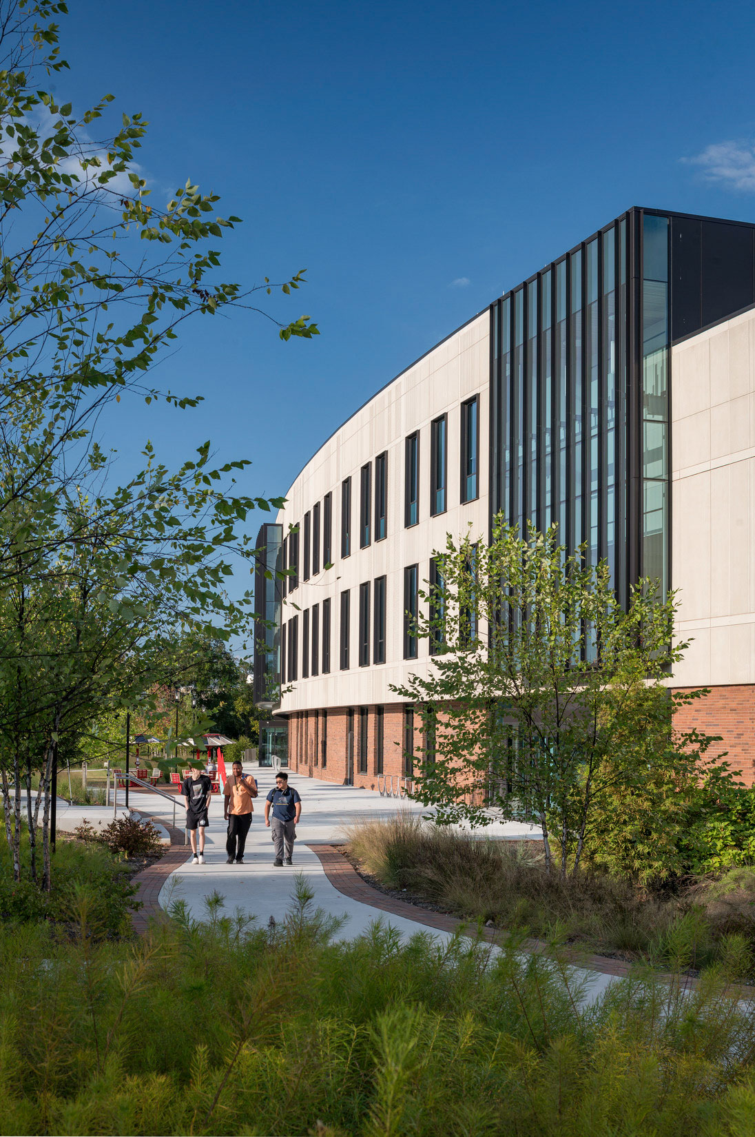 Exterior view with landscaped walkway and trees leading to modern building with glass and brick...
