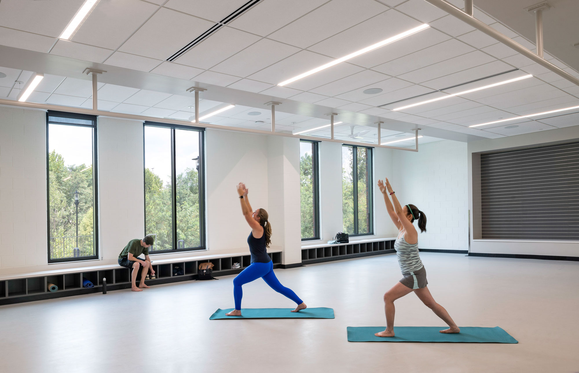 Exercise studio with large windows and two people practicing yoga poses on mats; bright, minimalist...