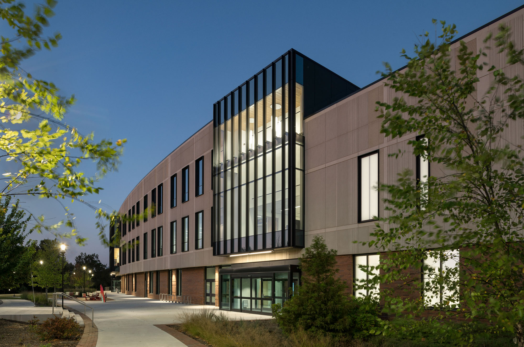 Evening view of building entrance with illuminated glass panels and landscaped walkway.