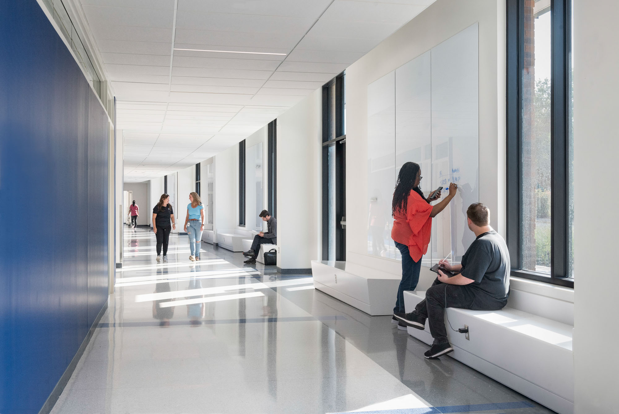 Bright hallway with white walls, large windows, and built-in benches; students walking and writing...