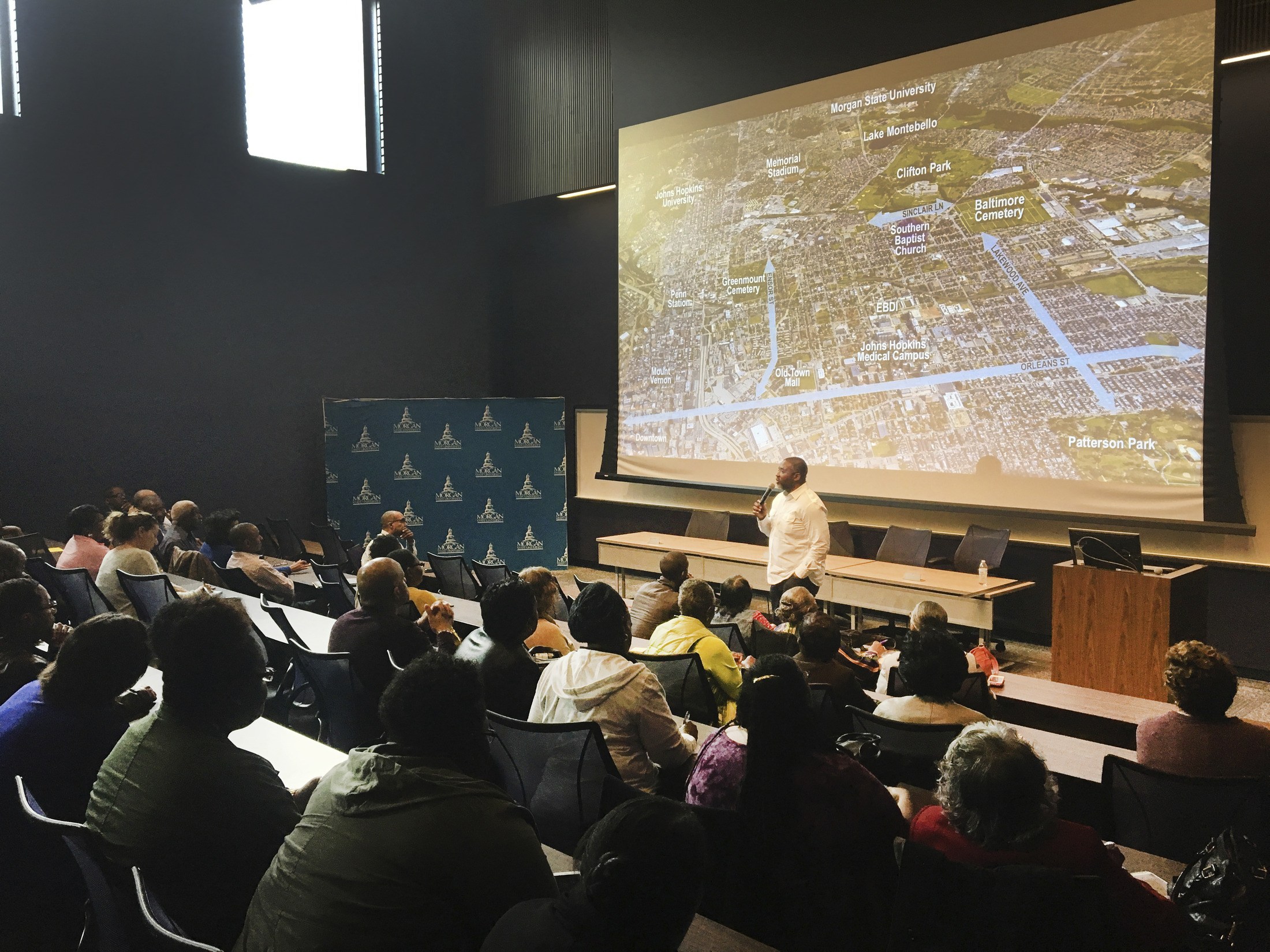 A person standing beneath a projection screen with a planning image on it addresses a crowd seated...