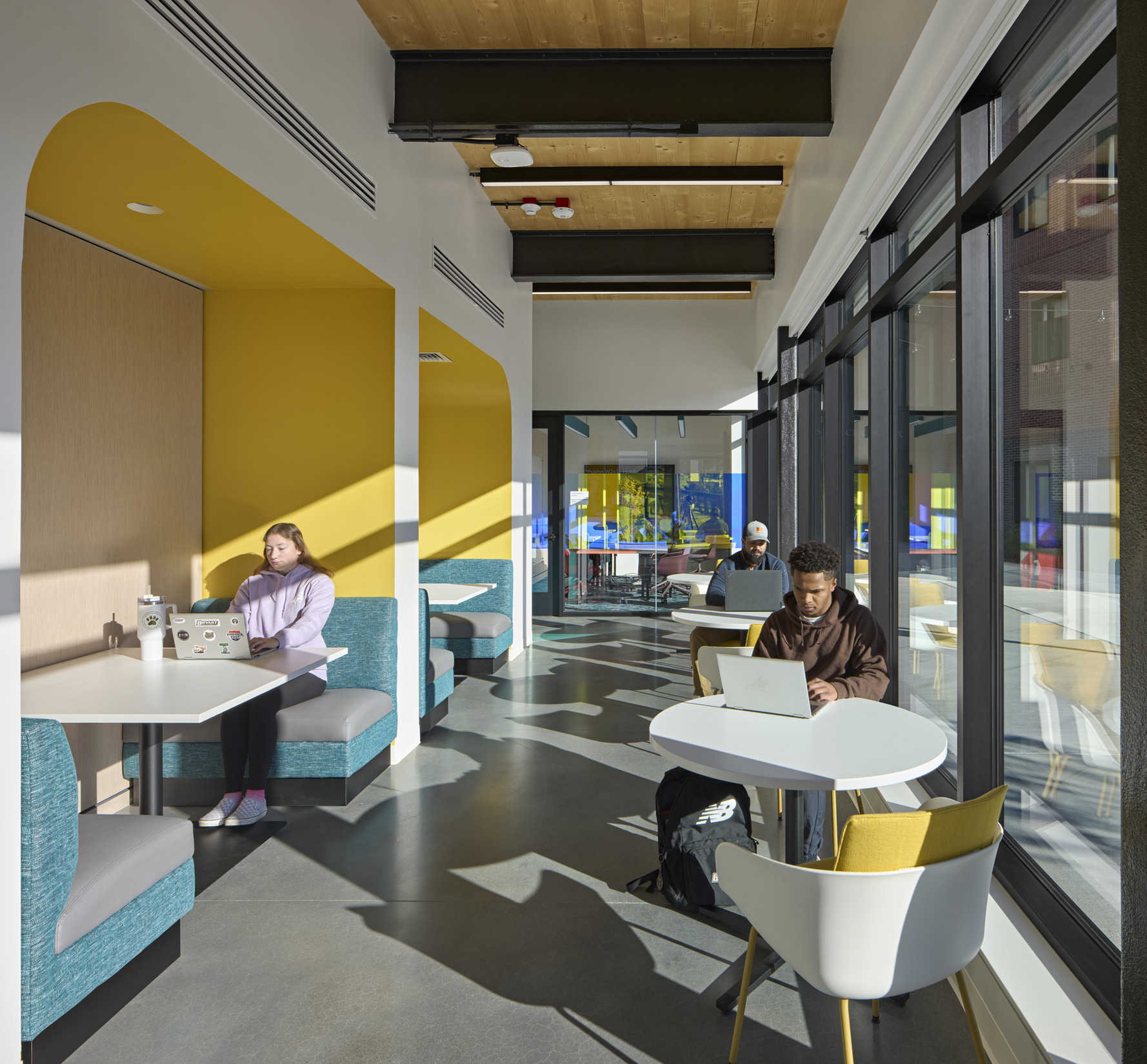 A hallway-style space with built-in booths featuring teal seating and white tables. Yellow accent...