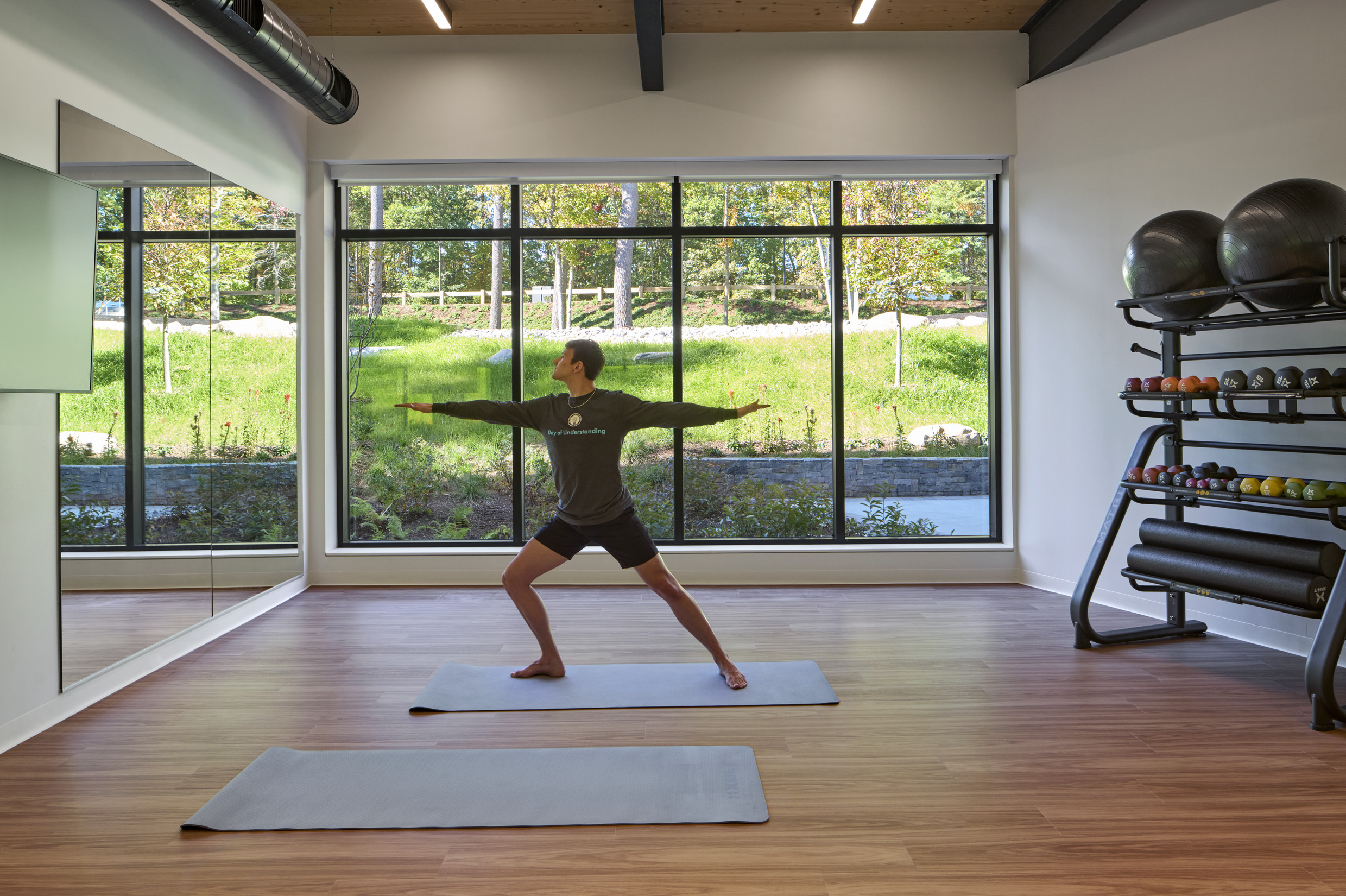 A bright fitness room with wood floors, two yoga mats, and a rack of weights and exercise balls....