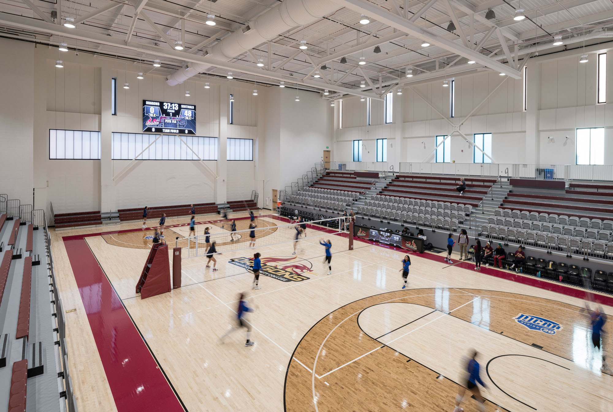 An indoor collegiate volleyball court with athletes practicing on a wood floor. Tiered bleachers...