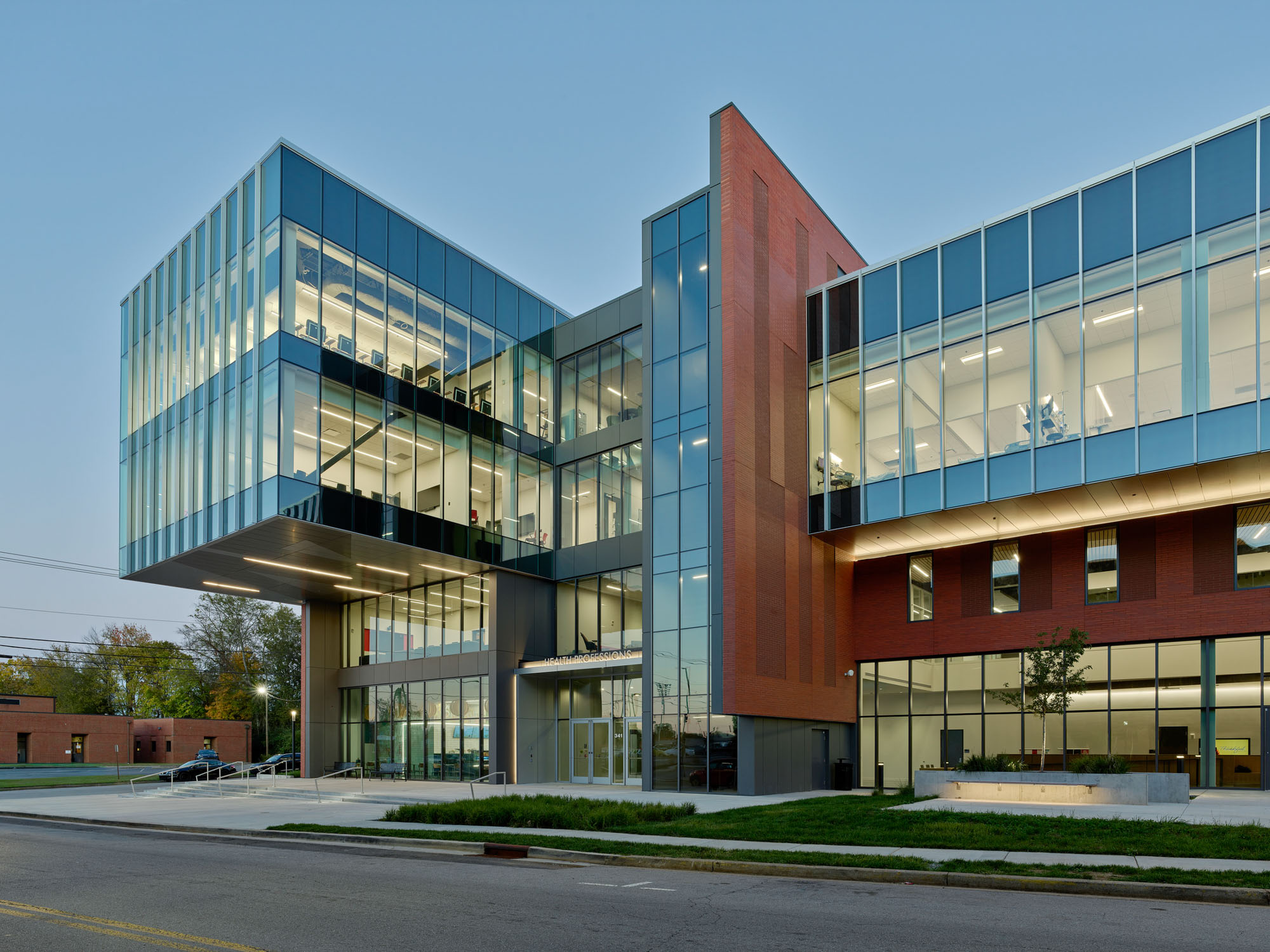 Contemporary glass-and-brick academic building illuminated at dusk, with a cantilevered upper level...
