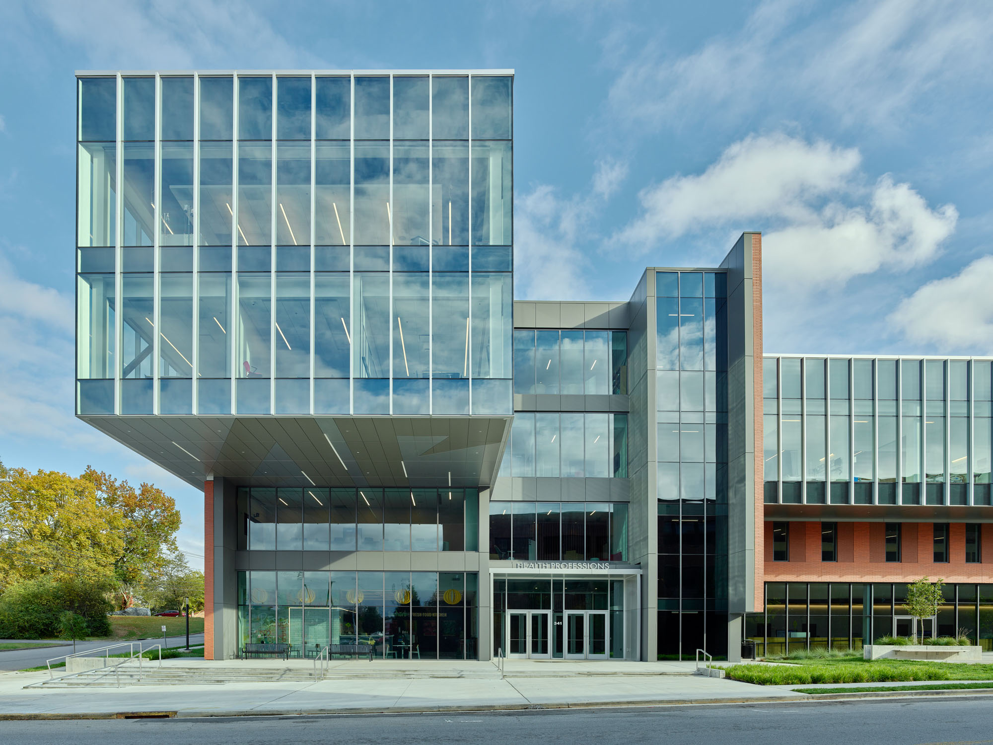 Three-story glass volume with vertical mullions projecting over the entrance, reflecting sky and...
