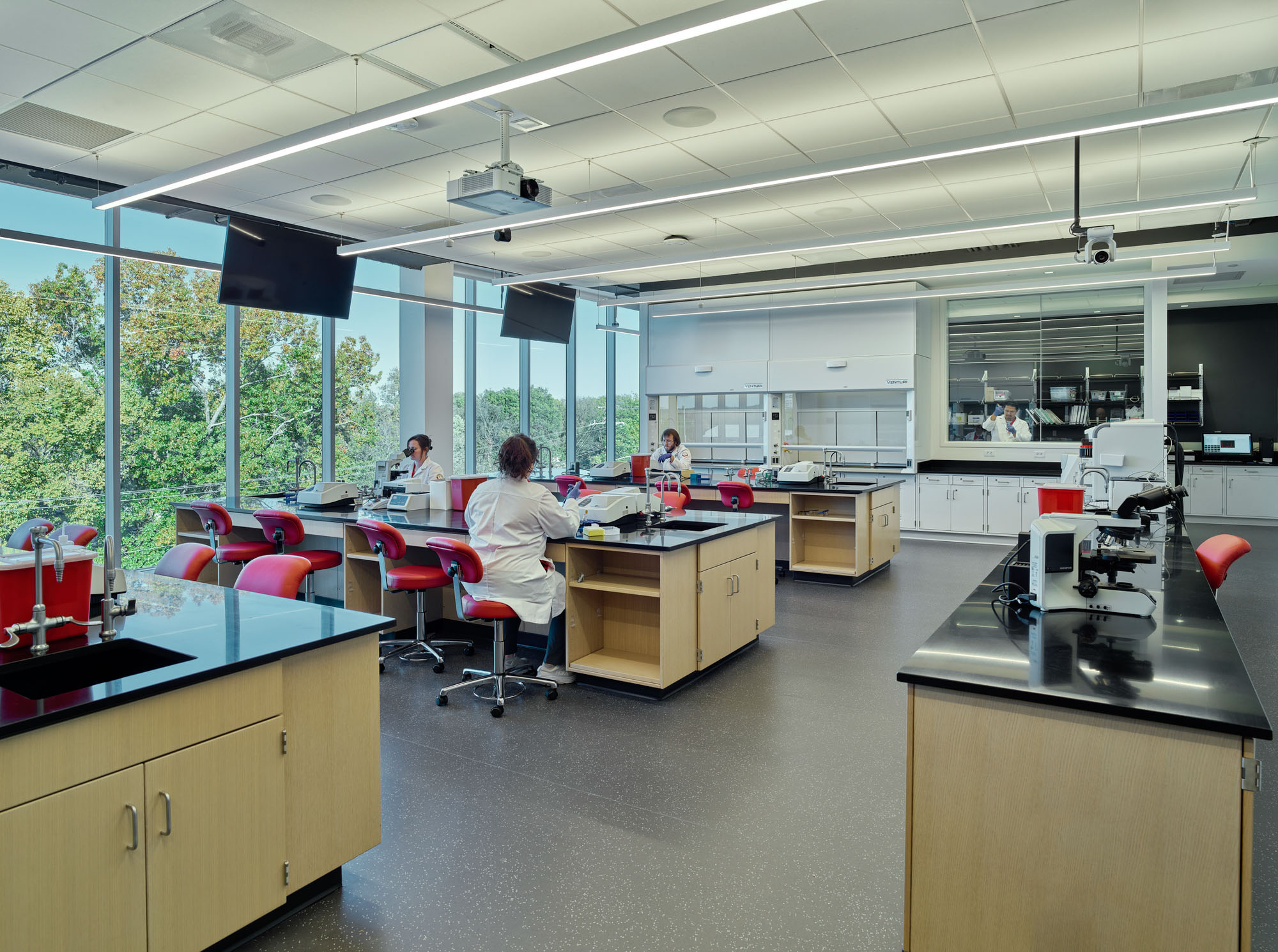Students in lab coats conduct experiments in a bright teaching laboratory with workstations, microscopes,...