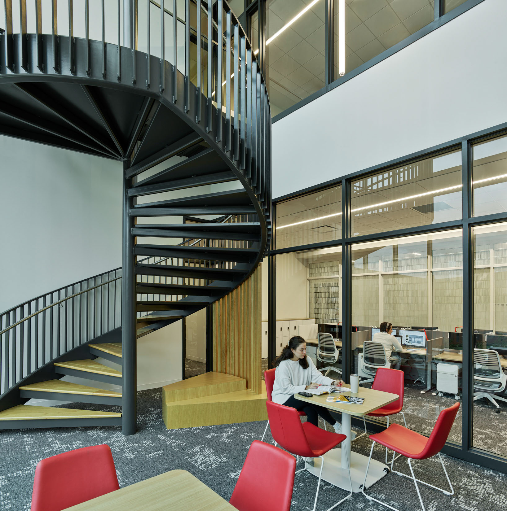 Black metal spiral stair adjacent to a glass-walled study room where students work at tables with...
