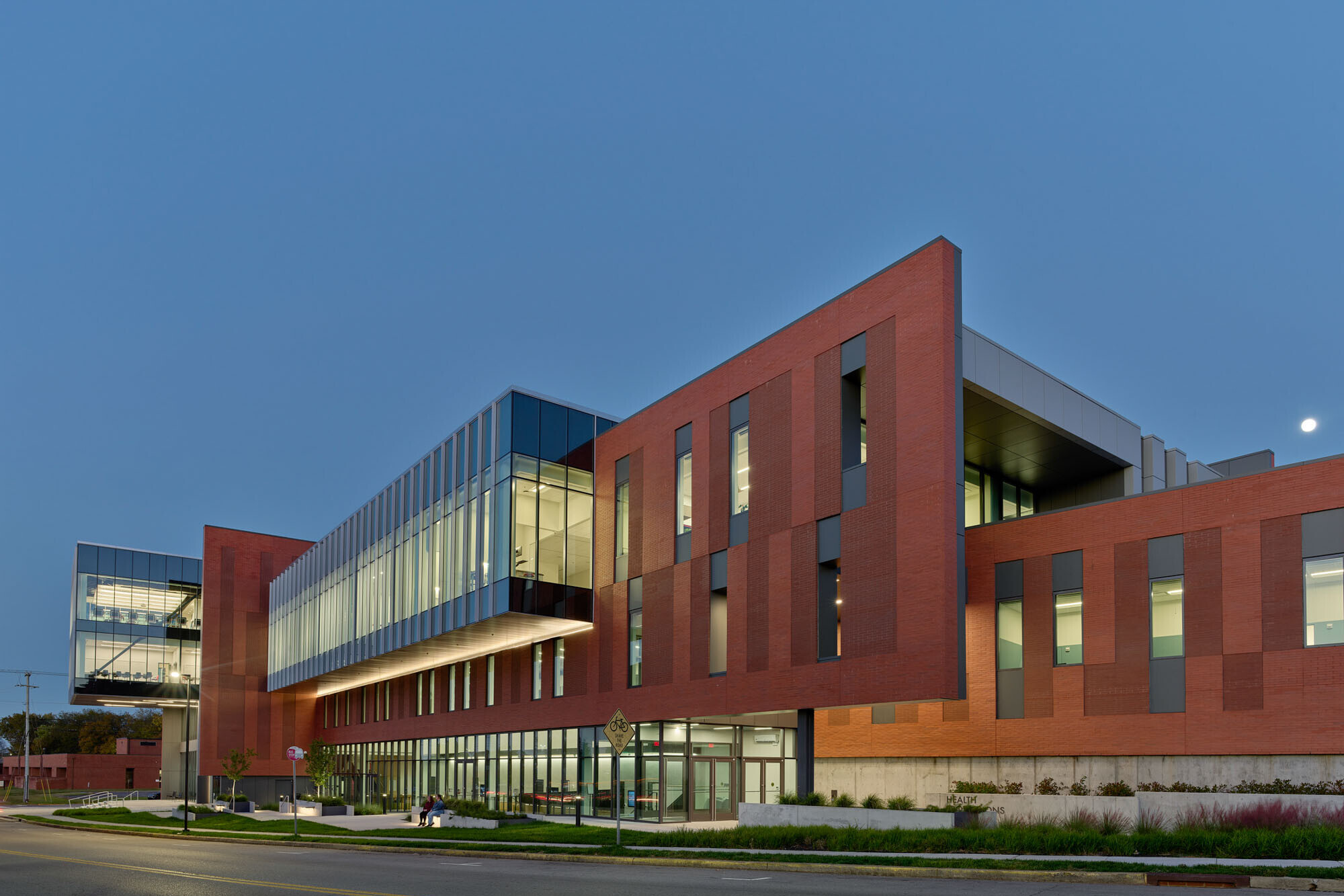 Health Professions Building at dusk featuring brick façades, illuminated glass volumes, and a cantilevered...
