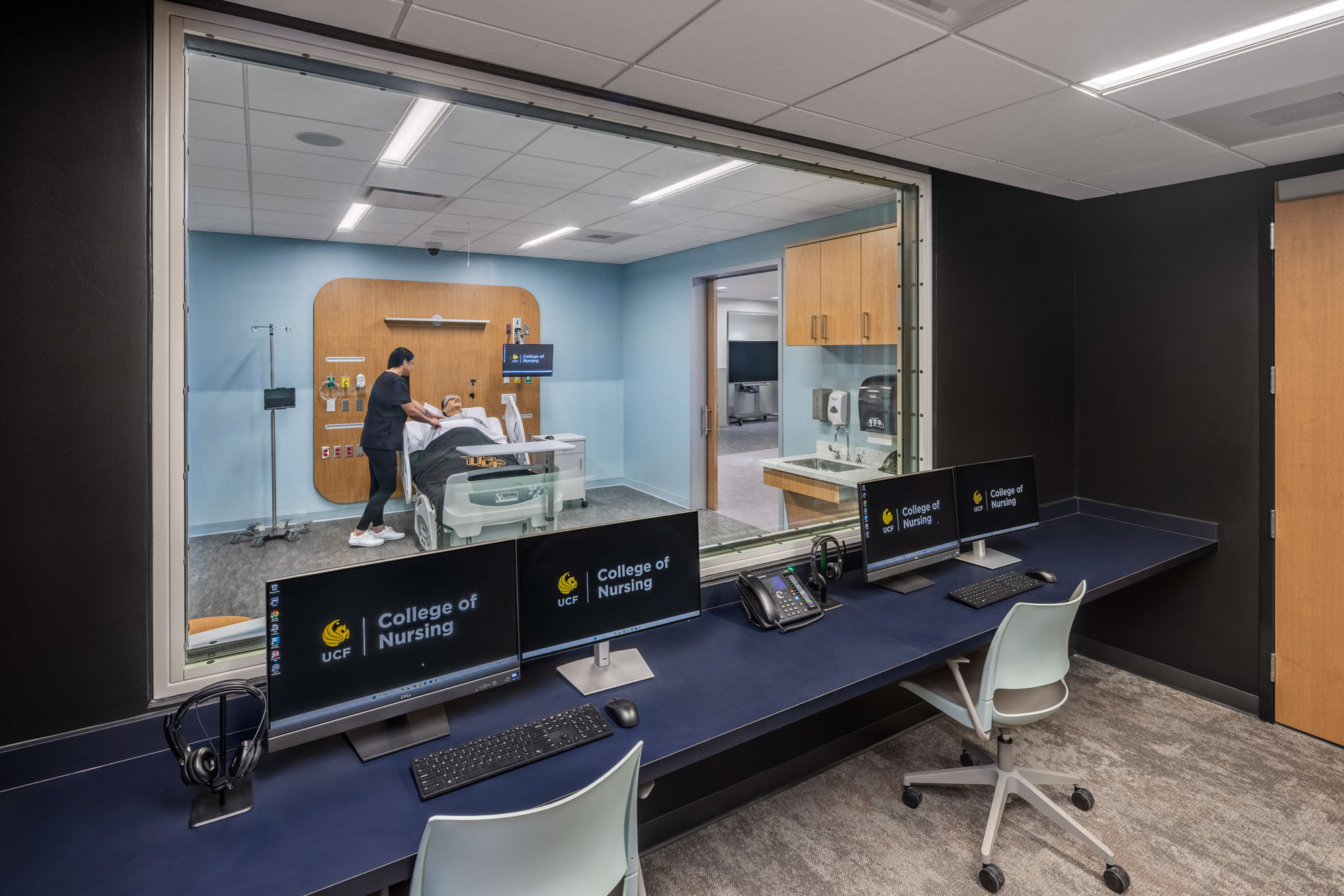 Simulation observation room with computer monitors overlooking a mock patient room through a large...