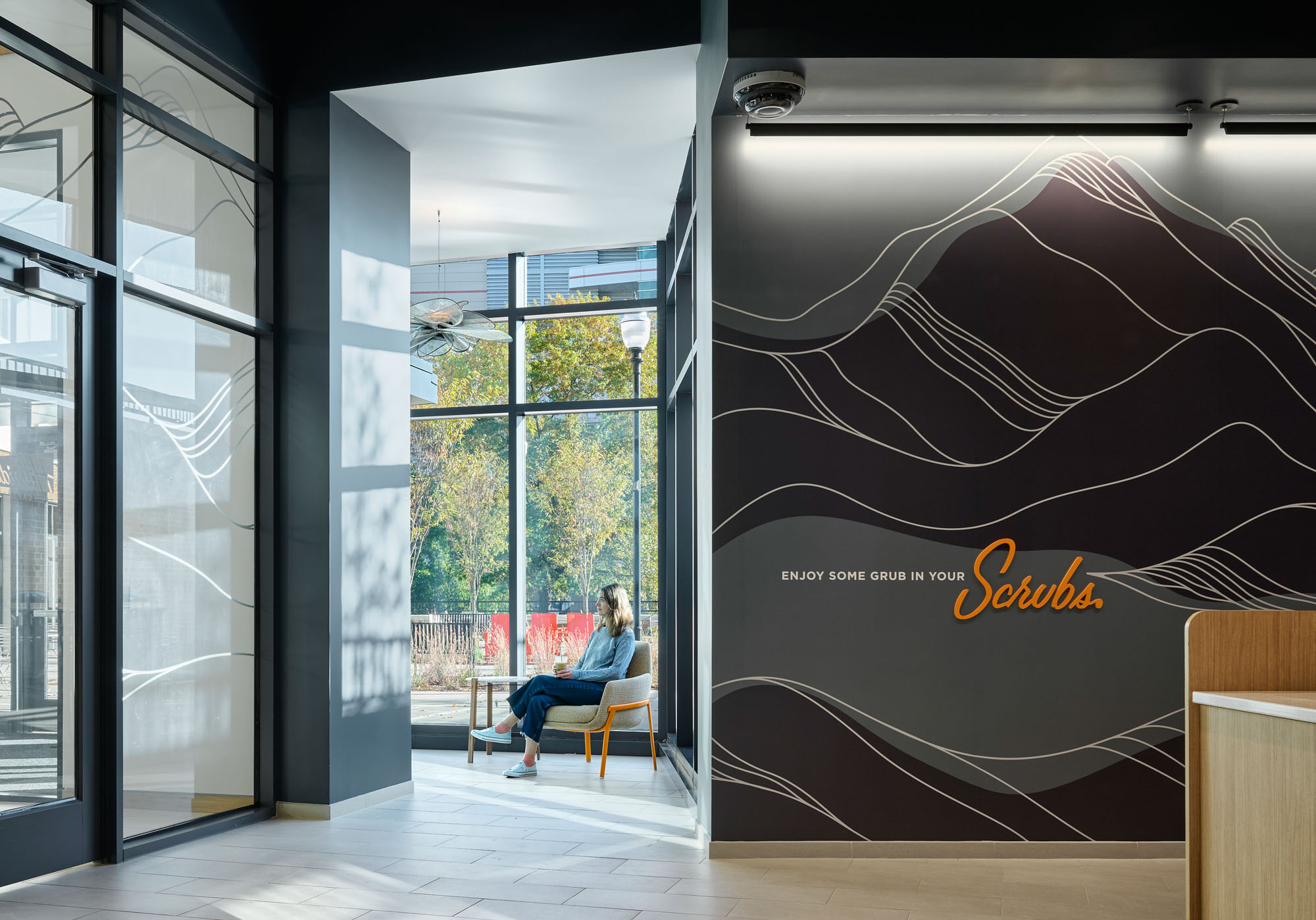 Interior café seating area with a student sitting near a floor-to-ceiling window, featuring graphic...