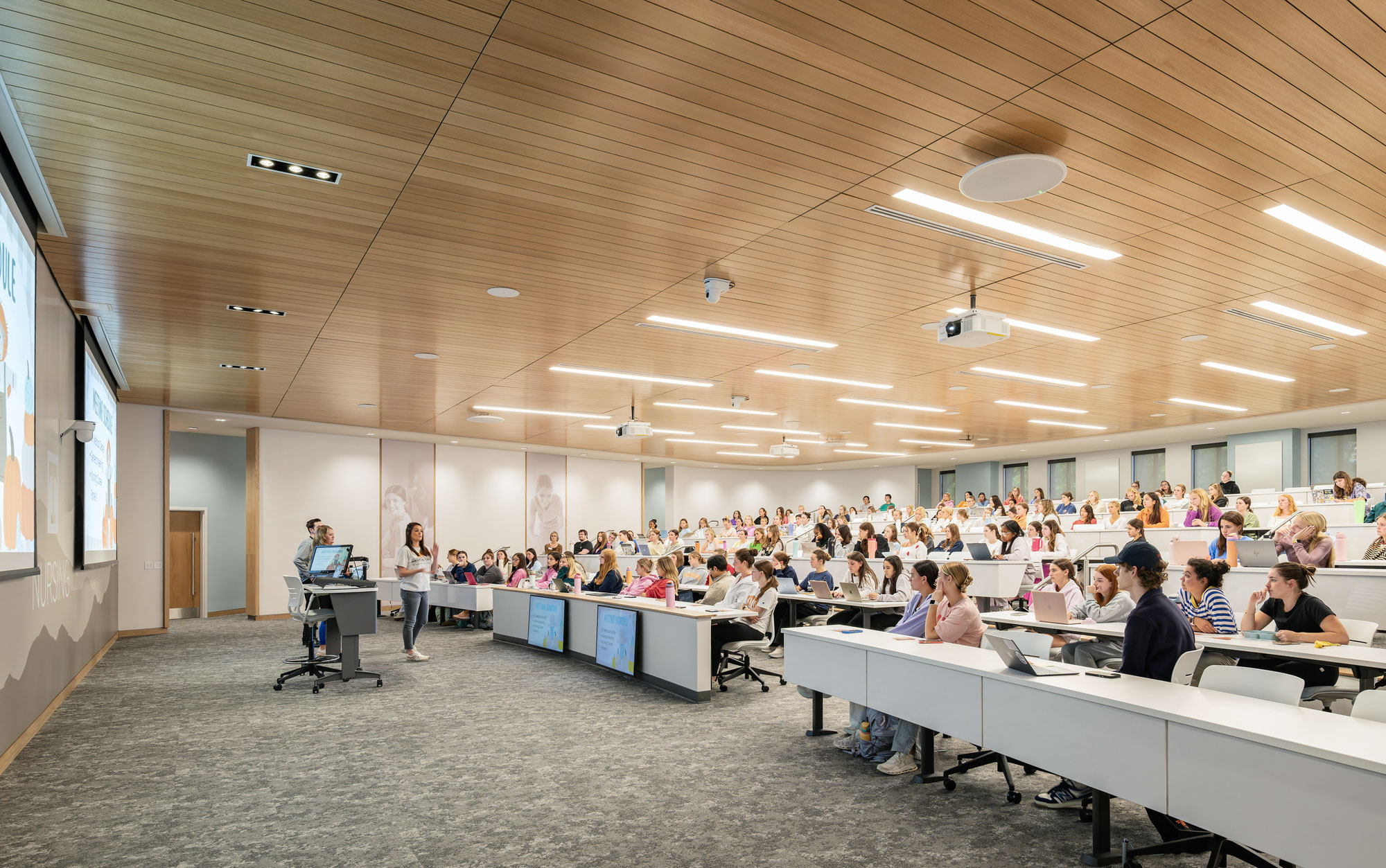 Tiered nursing classroom with students seated at desks, integrated technology, large projection...
