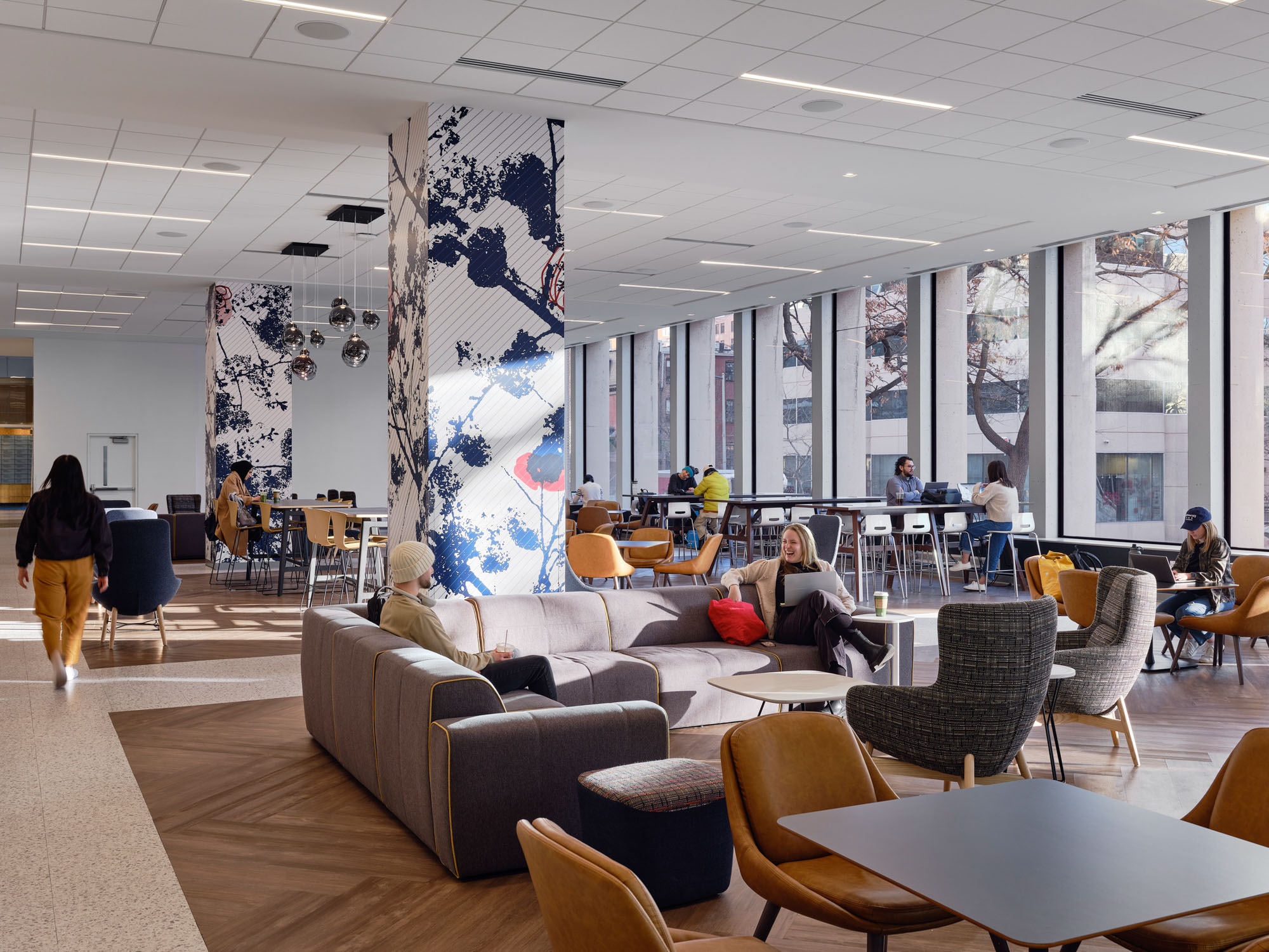 A university student lounge bathed in natural light from floor-to-ceiling windows features a mix...