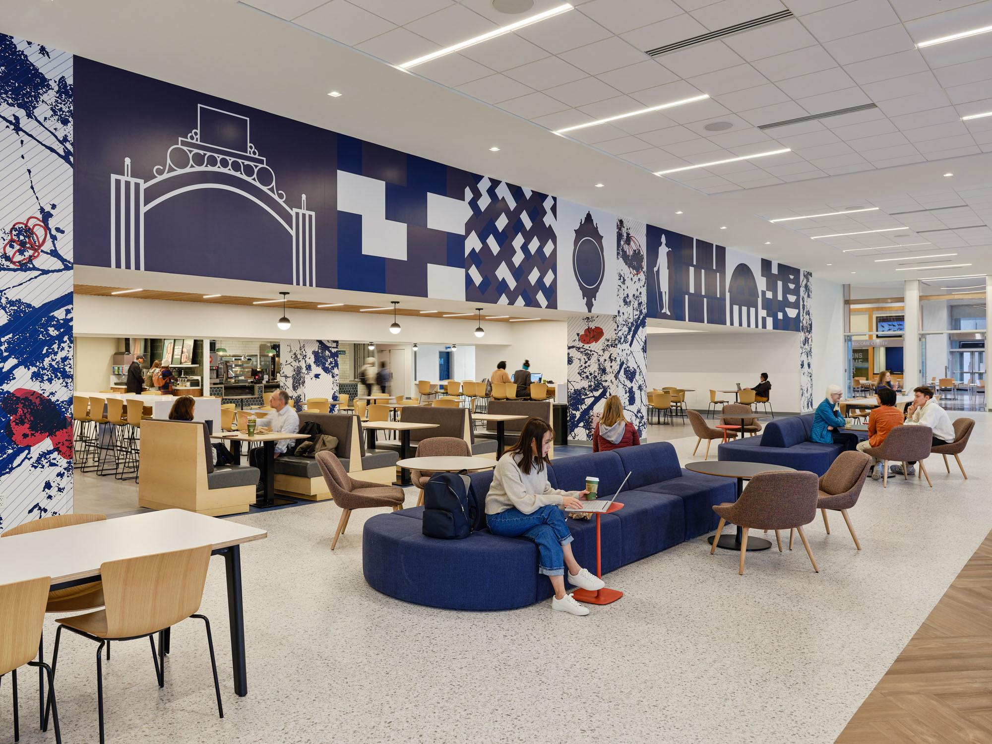 A wide view of a university dining commons shows a bold, large-scale mural spanning the upper wall,...
